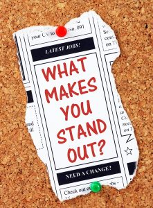 The question What Makes You Stand Out in red text on a newspaper clipping from the classified advertising section pinned to a cork notice board